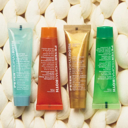 A set of four small Peter Thomas Roth skincare masks on a textured, off-white background. The tubes are in shades of light blue, orange, gold, and green. This mask kit is available from Skin & Glow